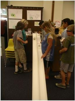 Photo shows students testing their model cars on two side-by-side tracks angled from a tabletop to the floor. 