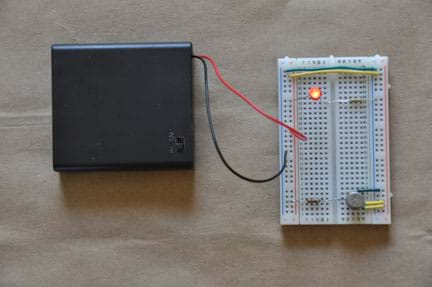 Photo shows a breadboard (circuit board) with the following connections: a yellow wire, which connects the positive terminals on either side of the board; a green wire, which connects the negative terminals on either side of the board; an LED, which is connected to the positive terminal on one side, and the center of the board on the other side; a resistor, which is connected to the same column as the LED in the center of the board, and the negative terminal on the other side; a VOC sensor; a battery pack. The LED is illuminated. 