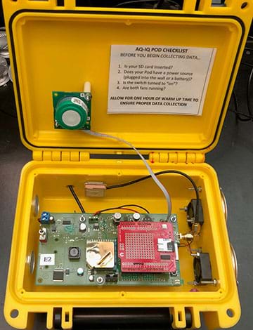 A photograph shows a Pod air quality monitor, which is a black briefcase-type container, shown with the lid open to reveal sensors and wiring. Nearby are example pollutant sources: rubbing alcohol, disposable lighters, super glue and votive (tea light) candles.