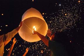 Photo shows people holding a domed lantern with a burning candle inside, and a dark sky filled with thousands more floating lanterns.