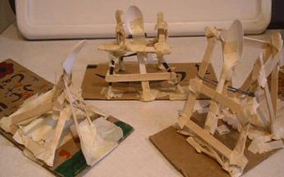 Photo shows three catapults made from wooden sticks, tape, rubber bands and plastic spoons, mounted to cardboard bases.
