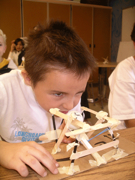 A photo shows a young student aiming a handmade Ping-Pong ball catapult made from wooden sticks, tape, rubber band and a plastic spoon.