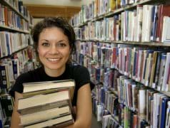 Photo shows a young girl holding a stack of books, surrounded by shelves full of books in a library. 