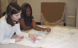 Two students at a table look at project solution ideas written on a large sheet of paper and applied sticky notes.