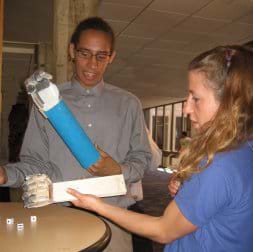 Photo shows two people at a table with forearm prostheses made from boards, eyelet screws, fishing line, plastic tubing and duct tape to make crude hands with fingers, tossing dice.