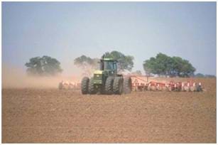 Photo shows a large green tractor with eight wheels pulling a very wide disc plow in a dusty field.