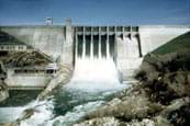 Photo shows water spilling over the top of a wide, straight concrete dam.