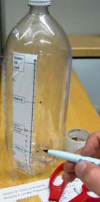 Photo shows a 2-liter clear plastic beverage bottle with a piece of marked paper taped on its side. A hand with a black marker makes two black circles on the plastic to the right of the Hole A and Hole B lines.