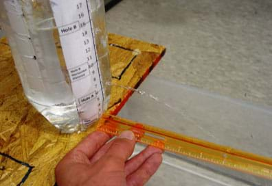 Photo shows a water-filled 2-liter clear plastic beverage bottle with a piece of marked paper taped on its side. A nearby hand holds a ruler from the base of the bottle to measure where the stream of water from the Hole A location lands.