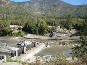 Photo from a riverside shows a concrete dam, half removed from blocking a river, with water flowing through the unobstructed side. 