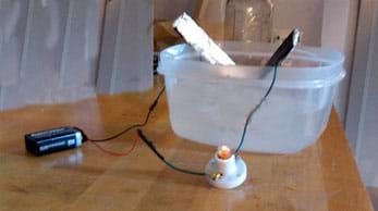 On a workbench, a 9-volt battery wired to a miniature light bulb socket and electrodes submerged in a tub of saltwater, with the light bulb on.