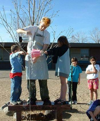 A photograph shows two students holding each dowel end of a waterwheels as a teacher helper pours water over the waterwheel fins.