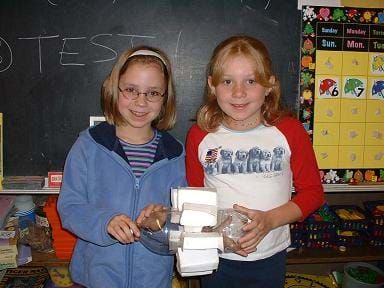 A photograph shows two girls holding a clear plastic two-liter botttle with box-shaped fins attached around its middle section.