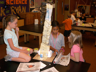 A photograph shows three students sitting around a tower made of recycled materials.