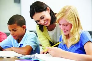 A photograph shows two students working on a writing assignment with a teacher nearby reviewing their work.