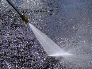 A photograph shows a hand directing a high-pressure stream of water from a hose onto an asphalt road surface. It's a Honda GX160 5.5 HP. pressure washer in action.