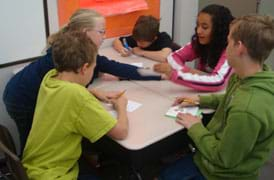 A photograph shows five youngsters around a table, busy writing and passing papers to each other.