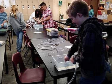 The image shows a photograph of a student testing his design in a classroom, while other students test their designs in the background.