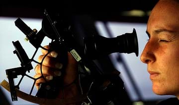 A photograph shows a side view of a woman's head as she looks through a marine sextant to determine celestial navigation aboard a ship.