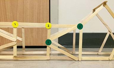 A photograph shows the side view of a bridge truss structure made from wooden craft sticks hot glued together. It looks like a geometric web of sticks connected together to form various polygon shapes. Two small round yellow stickers, marked “1” and “2,” are placed at two adjacent corners of a square truss component composed of four sticks. Nearby, two small round green stickers, marked “1” and “2,” are attached to two adjacent corners of a triangle truss component. The stickers identify angles will be measured before and after load testing to check for deformation.