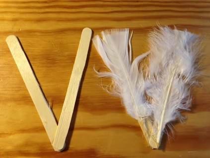 A close up photograph shows two craft sticks glued together at a 40° angle, which looks like a "V" (left) and another two craft sticks prepared in the same way but covered with white feathers (right).