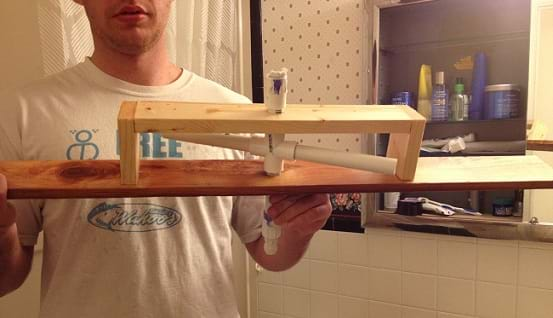 A young man holds up a PVC piping pump shaped like a plus sign mounted in a wooden frame.