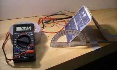 Photo shows a tabletop with a handheld multimeter connected by red and black wires to a PV panel that is tilted at an angle, being measured by a protractor.