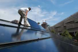 Two men standing on an angled roof lay a shiny blue-black panel on its surface.