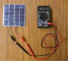 Photo from above a tabletop shows red and black wires from a small PV panel connected to red and black leads from a digital multimeter.