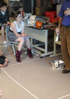 Photo shows students gathered outside a taped off floor area, ~ 1 x 1 meter in size, inside which a lamp is clamped on a desk ~.6 meters above the floor, facing down on a solar panel and foil contraption on the floor.