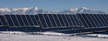 Four tilted rows of black panels mounted on the snowy ground under a blue sky with snowcapped mountains in the distance.