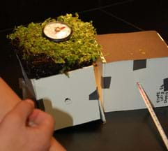 Photo shows two small cube structures made of taped-together foam core board. One rooftop has a square of sod with a round thermometer sitting on it. A long thermometer pokes into the interior of the other structure.