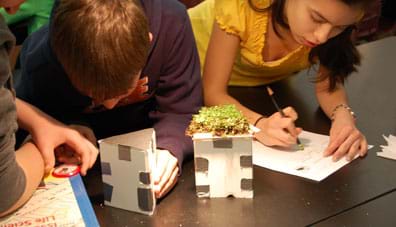 Photo shows three children at a table observing, reading and recording the temperatures of two small buildings made of foam core board and duct tape.
