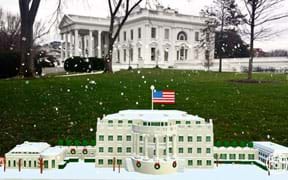 A photograph shows the U.S. president’s residence, the Whitehouse, surrounded by green lawn and trees, with snowflakes falling. In the foreground is a small scale model of the same building.