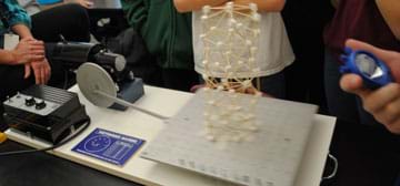 Students use a stopwatch as a tall toothpick and marshmallow structure jiggles on a tabletop platform.