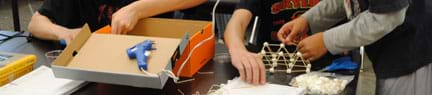 Three teens at a table with a cardboard box, glue gun and truss-like structure of toothpicks and mini marshmallows.