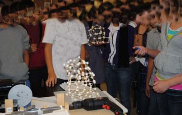 Photo shows many 8th grade students around a table, one holding a stopwatch, watching a marshmallow and toothpick structure move on an electronic mini shake table.