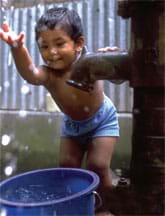 Photo shows a boy splashing water from a bucket under a steel water pump.