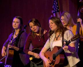 Four young girls play stringed instruments on a stage.