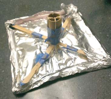 A photograph shows a flat square of aluminum foil with its edges wrapped around a square frame made from four plastic drinking draws and cross-bracing through the middle and into the four corners from taped-together wooden Popsicle sticks. A cardboard tube is secured vertically with rubber bands and tape where the sticks cross in the center.