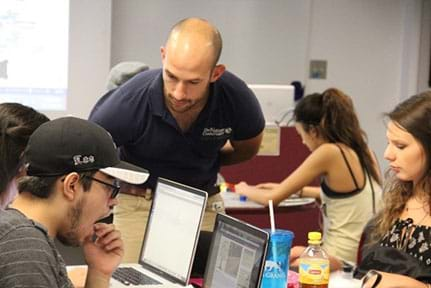 A photograph shows an instructor helping students as they work on computers.