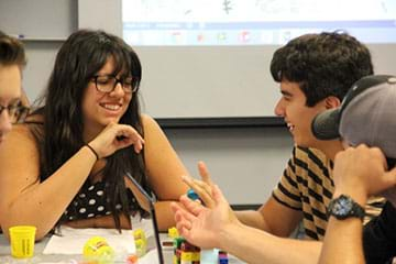 A photograph shows teenagers sitting at a table scattered with small colorful blocks, yellow clay and paper.