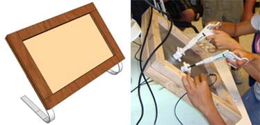 Two images: (left) Drawing shows a wooden frame around a latex rubber sheet with curved aluminum strips as support legs.(right) Photo shows two pairs of hands holding laparoscopic tools and a small digital camera, all inserted through slits in the framed rubber sheet.