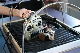 Photo shows a hand holding a transmitter next to a small three-wheeled electronic contraption on a surface of plump black tubes.