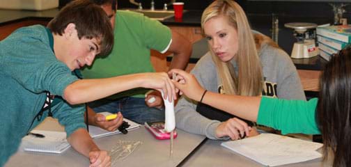 Photo shows four teens at a table with one person holding a ruler vertically with one end touching the table, another holding a cylindrically shaped piece of silly putty next to the ruler, another using a stopwatch for timing, and another ready to record measurements as they all watch the silly putty stretch towards the table.