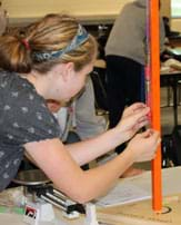 Photo shows a girl pulling on a spring scale that is hanging from a ring stand on a table. In turn, the spring scale is attached to a paper clip hook, small balloon and another paper clip hook that hang from the ring stand. Pulling the spring scale stretches the balloon.