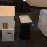 A photograph shows a small-sized foamcore board model house resting on an overturned cardboard box. On the right, a box fan blows air over a tray of ice toward the model house. 