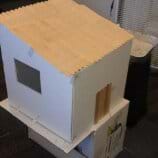 A photograph shows a small-sized model house made of foamcore board with a roof made of wooden craft sticks. The house is positioned on top of a cardboard box.