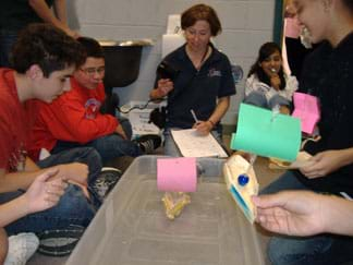 Holding a hair dryer towards a model ship in a pool of water, a teacher leads the race course challenge with students cheering on the sides.