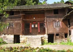 Photo shows the front view of a two-story wooden house raised on a stone foundation with a peaked tile roof, four windows and a centered double front door.